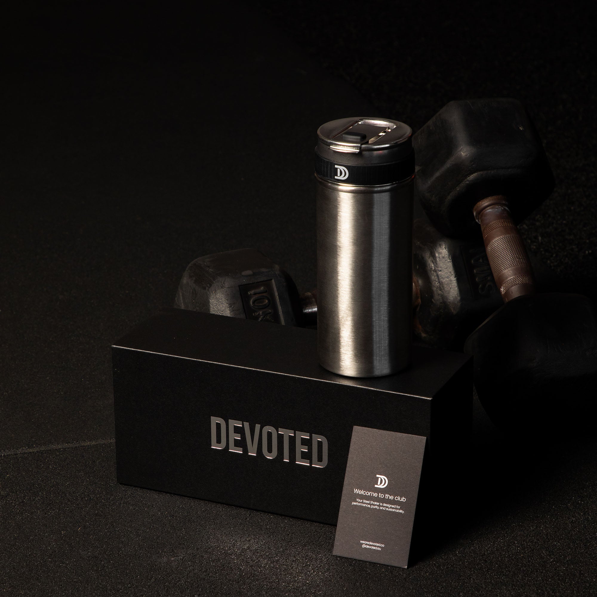 Devoted Steel Shaker silver stainless steel tumbler with black lid placed on black box against dumbbells