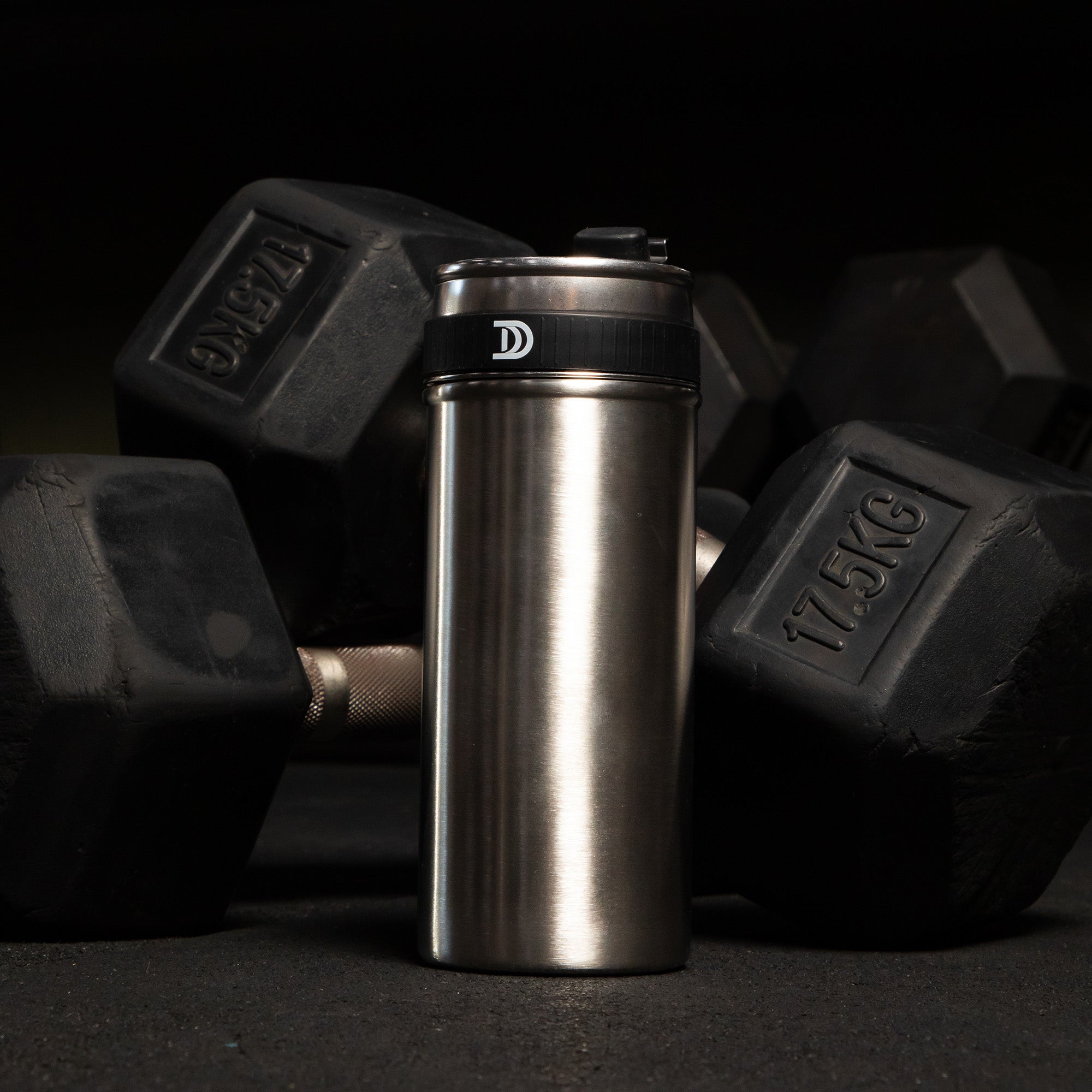 Devoted Steel Shaker with black lid surrounded by black dumbbells on a dark gym background 100% plastic-free stainless steel bottle